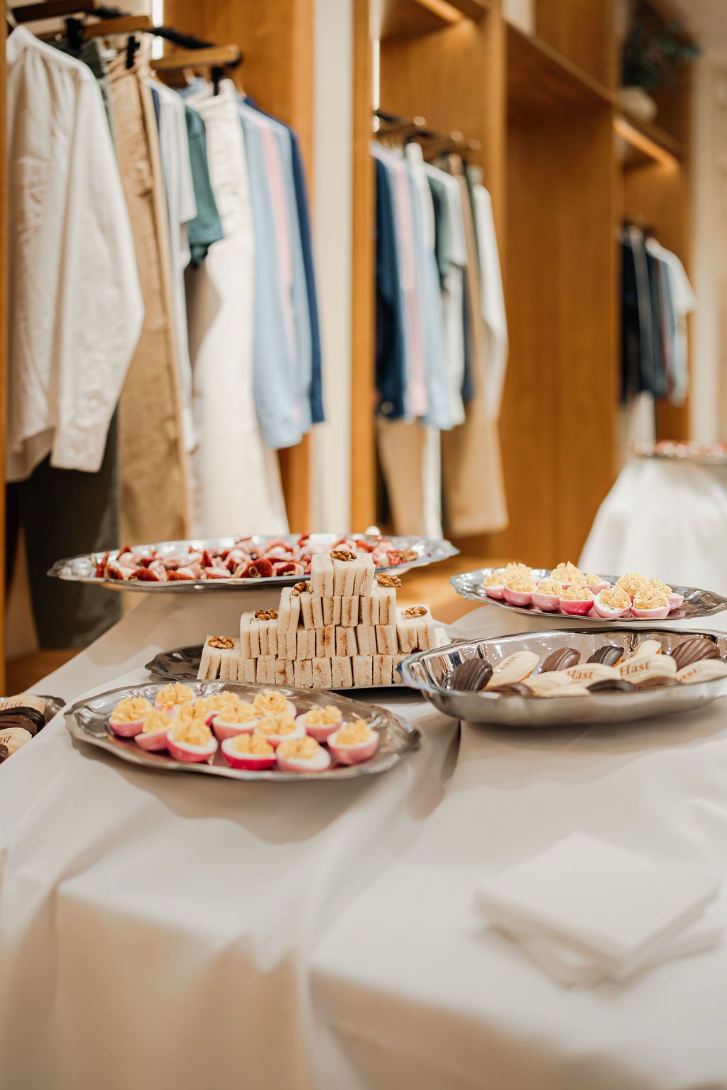 Table with plates of assorted appetizers including deviled eggs, small sandwiches, and chocolates in a room with hanging clothes.