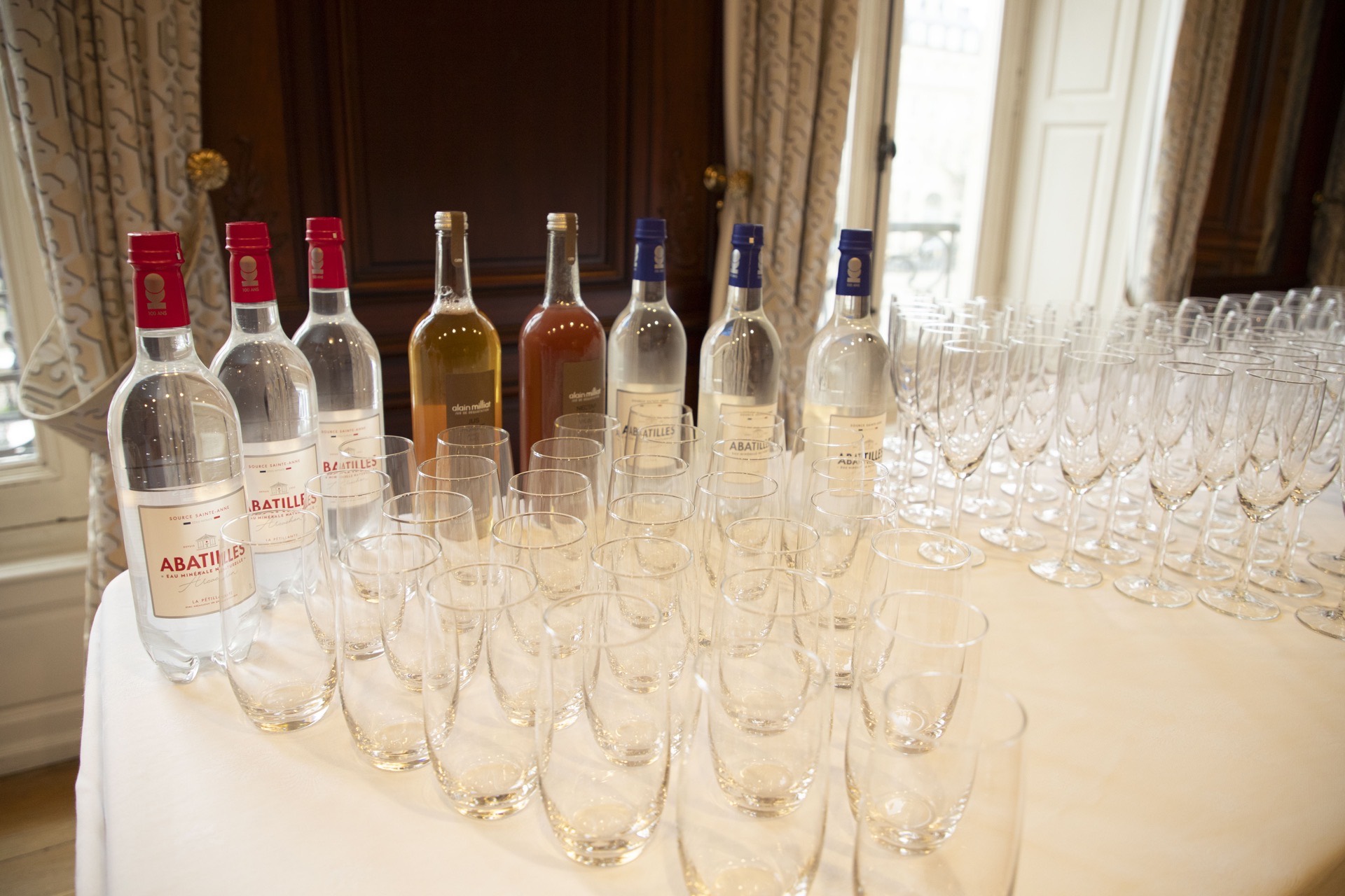 Bouteilles d'eau minérale et de jus avec rangées de verres vides sur une table blanche dans une salle élégante.