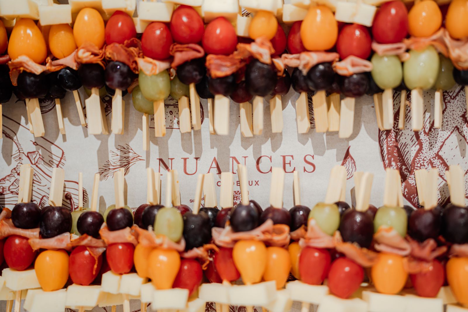 Brochettes colorées avec cubes de fromage, raisins rouges et verts, tomates cerises et tranches de jambon disposées sur un plateau marqué « Nuances ».