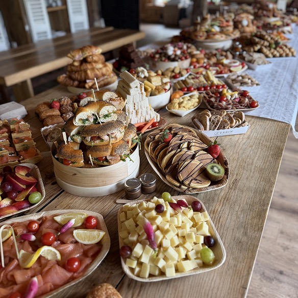 Table en bois garnie d'un buffet varié avec des sandwichs, plateaux de fromage, fruits, saumon fumé, viennoiseries et pancakes décorés de fruits.