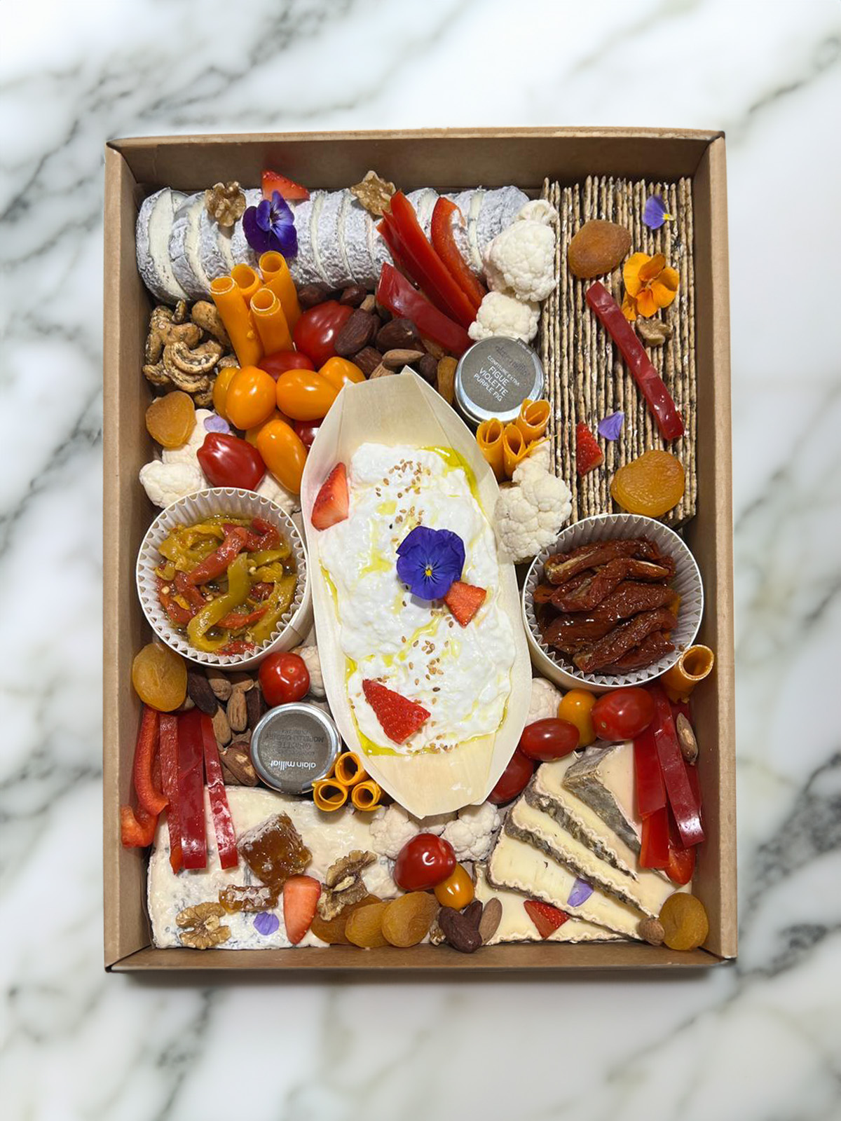 Plateau de fromages variés, légumes frais, noix, abricots secs, tomates cerises, crackers et fleurs comestibles dans une boîte en carton.