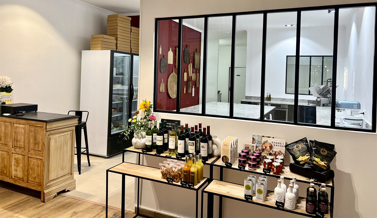 Intérieur d'une boutique élégante avec comptoir en bois, bouteilles de vin, produits gourmets et fenêtre donnant sur une cuisine moderne.