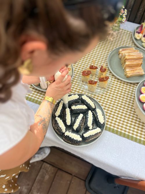 Une femme décore des petits pains noirs avec une crème blanche à l'aide d'une poche à douille sur une table à carreaux avec divers amuse-gueules.
