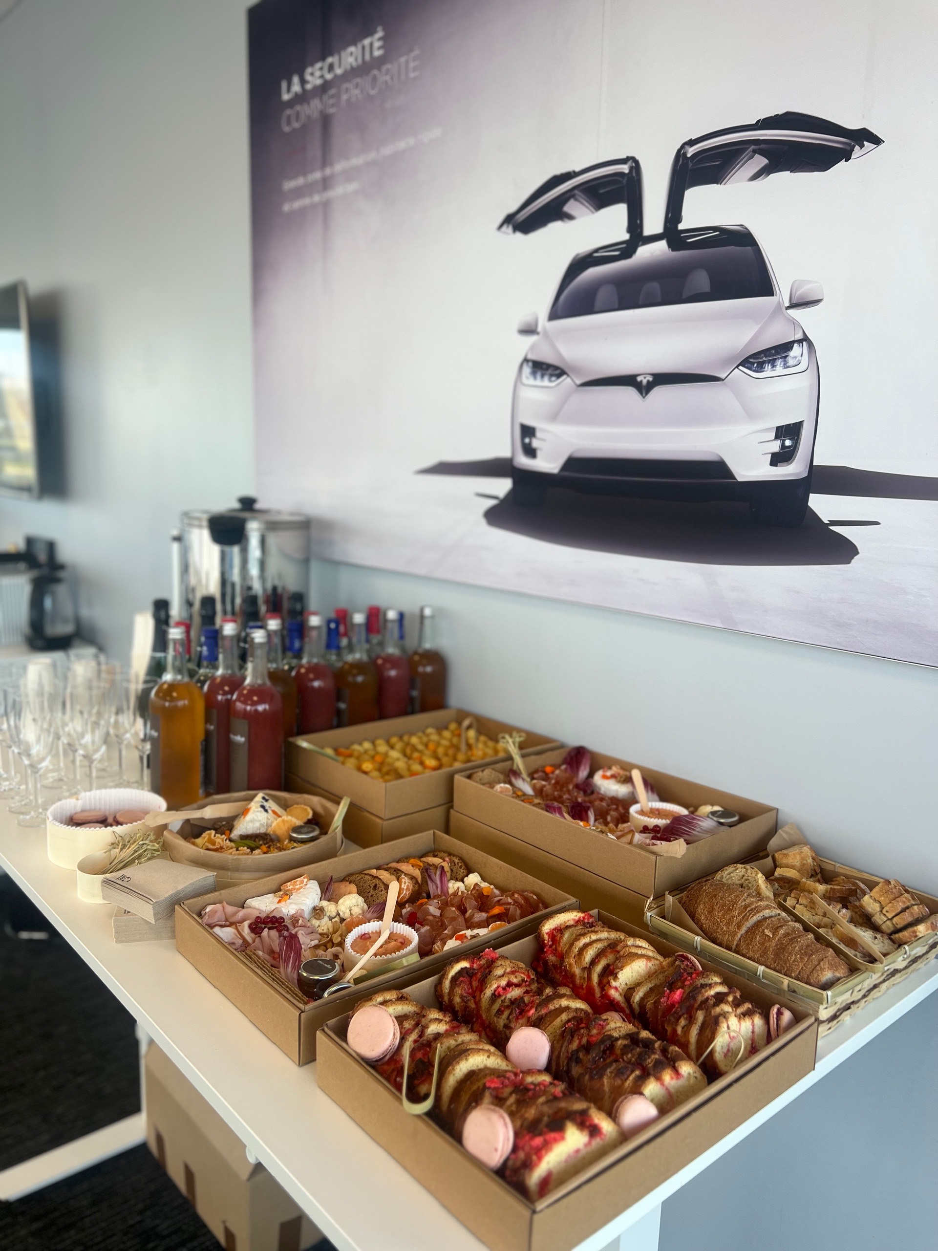 Buffet de nourriture et boissons sur une table blanche avec une affiche de voiture électrique Tesla blanche au mur.