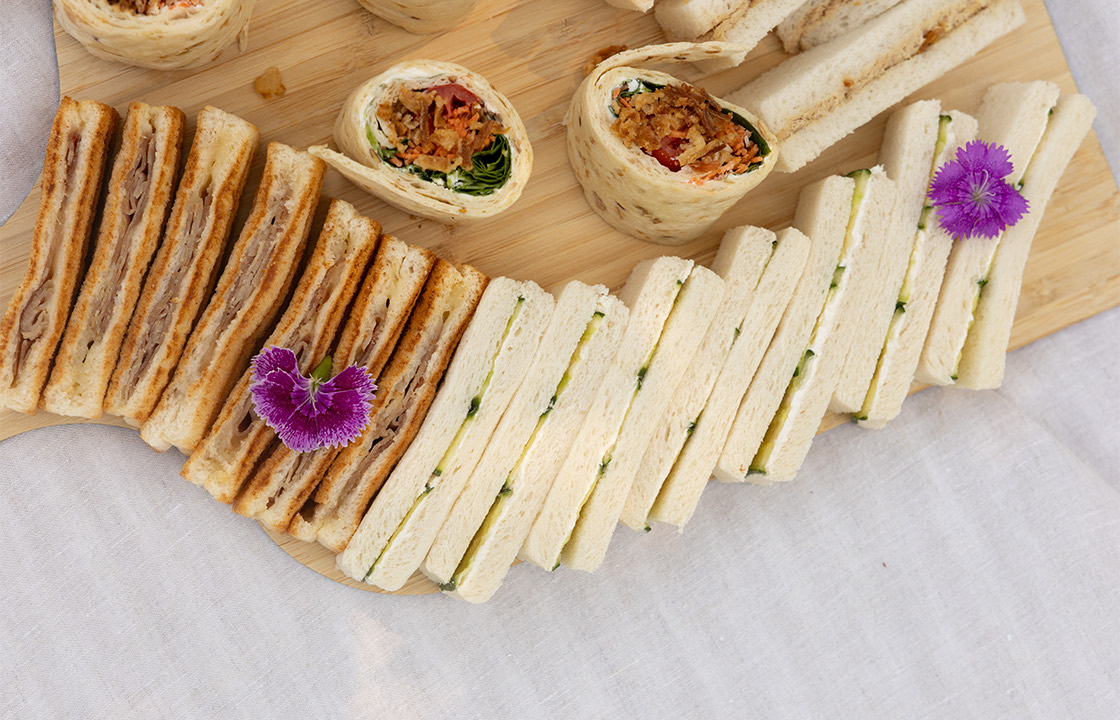 Plateau de sandwiches variés comprenant des sandwiches grillés au jambon, des sandwiches à la crème et concombre, et des wraps garnis de légumes et d'oignons frits, décorés de fleurs comestibles violettes.
