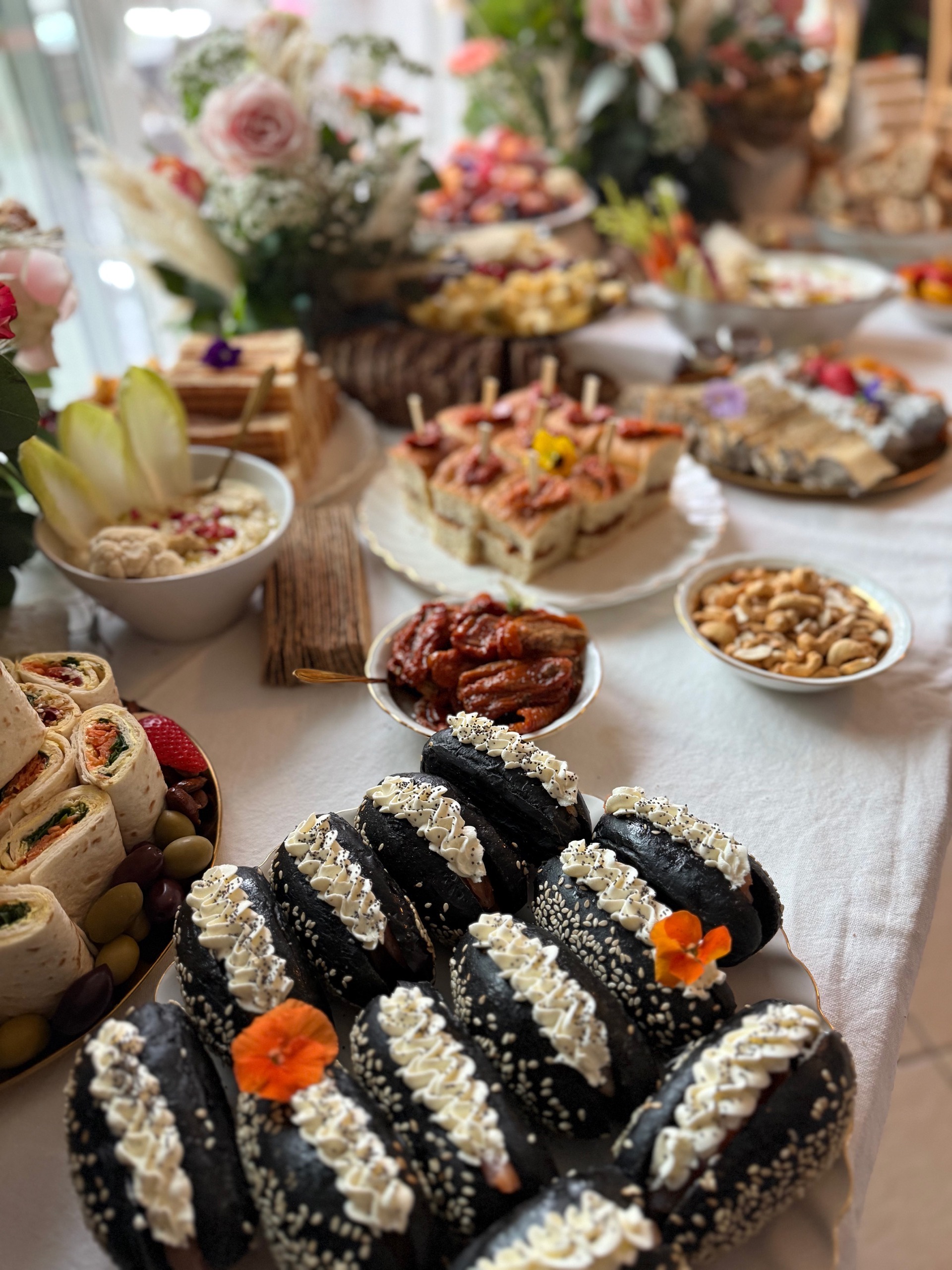 Buffet table with assorted gourmet finger foods including black sesame seed buns with white cream, wraps, nuts, dried fruits, and decorative flowers.