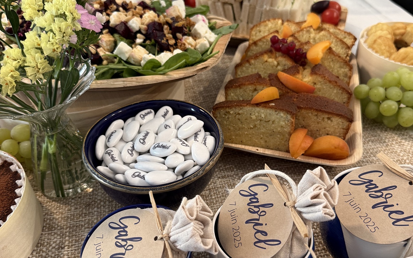 Table décorée avec fleurs, salade aux noix et fromage, tranches de gâteau, raisins verts, et dragées blanches personnalisées avec le nom Gabriel.