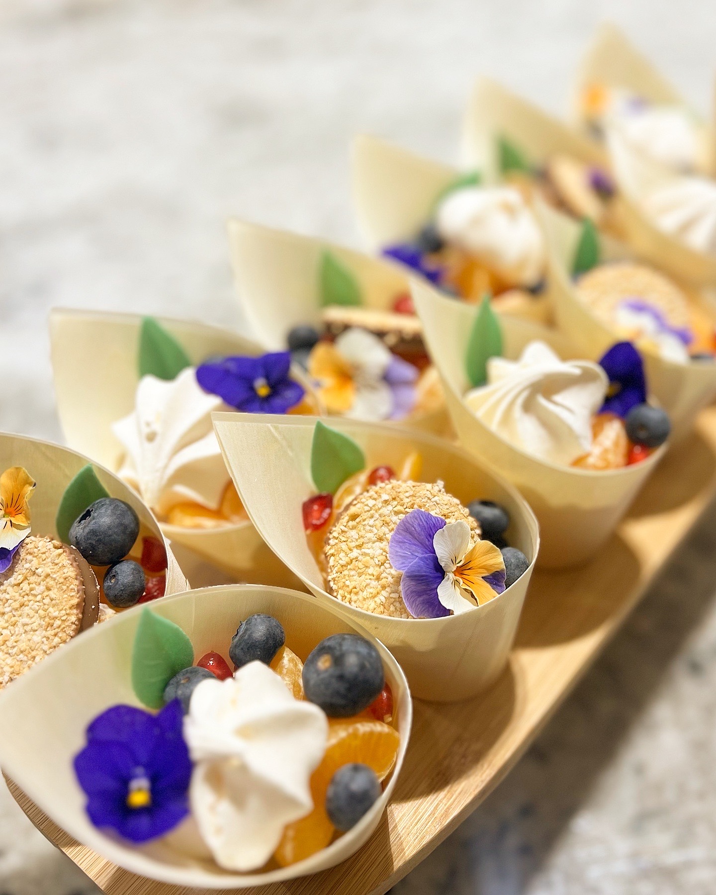 Petites coupes de dessert décorées de meringue, myrtilles, tranches de mandarine, fleurs comestibles et biscuits croustillants sur un plateau en bois.
