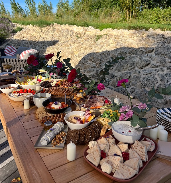 Buffet en plein air sur table en bois, avec pains, fromages, charcuterie, dips, fruits secs et fleurs décoratives.